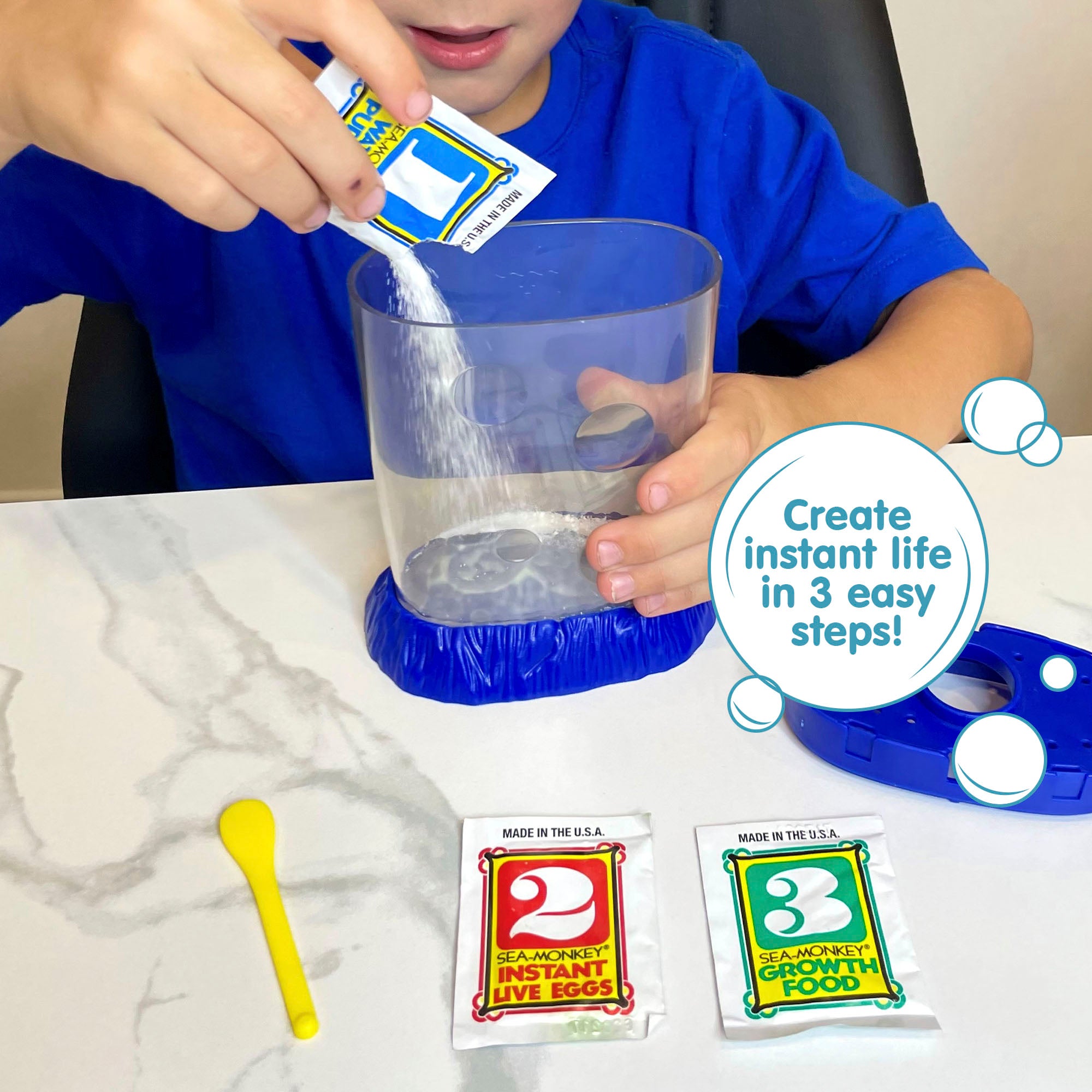 A child wearing a blue shirt is holding the Magiquarium with one hand, and pouring powder from a small packet labled with the number 1 with the other hand. The lid of the aquarium, a packet with the number 2, a packet with the number 3, and a yellow plastic spoon all lay on the surface in front of the child.